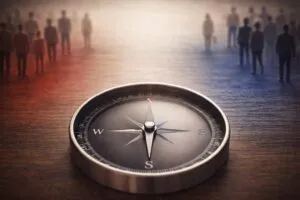 A compass lies on a wooden surface, its needle pointing north. In the blurred background, silhouettes of people stand divided
