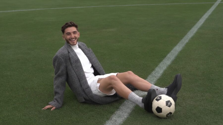 A person in a gray coat and white shorts sits smiling on a grassy soccer field