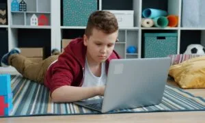 Boy in a red hoodie lies on a striped rug using a laptop