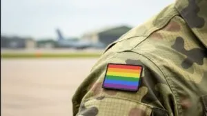 A person in camouflage military uniform is shown from the back, with a rainbow pride flag patch on the shoulder