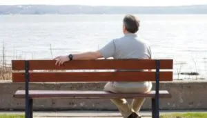 A man in a gray shirt sits on a wooden bench, facing a calm lake