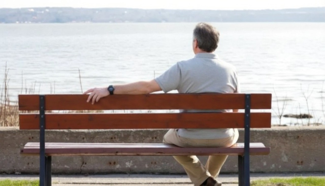 A man in a gray shirt sits on a wooden bench, facing a calm lake