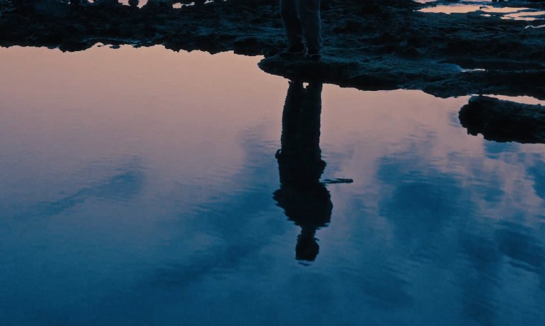 Silhouette of a person standing on rocky terrain, reflected in a calm body of water during sunset