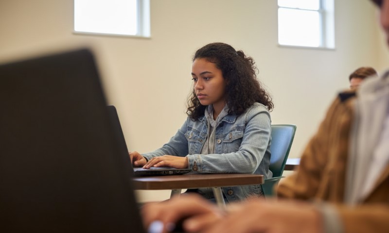 A focused student in a denim jacket sits at a desk, typing on a laptop
