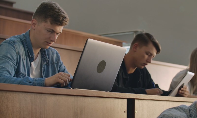 Two students in a lecture hall focus on laptops, seated at wooden desks
