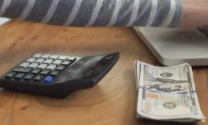 A close-up shows a hand on a laptop, a stack of hundred-dollar bills, and a black calculator on a wooden desk