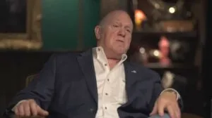 Tom Homan sits in a blue suit and white shirt sits in a dimly lit room