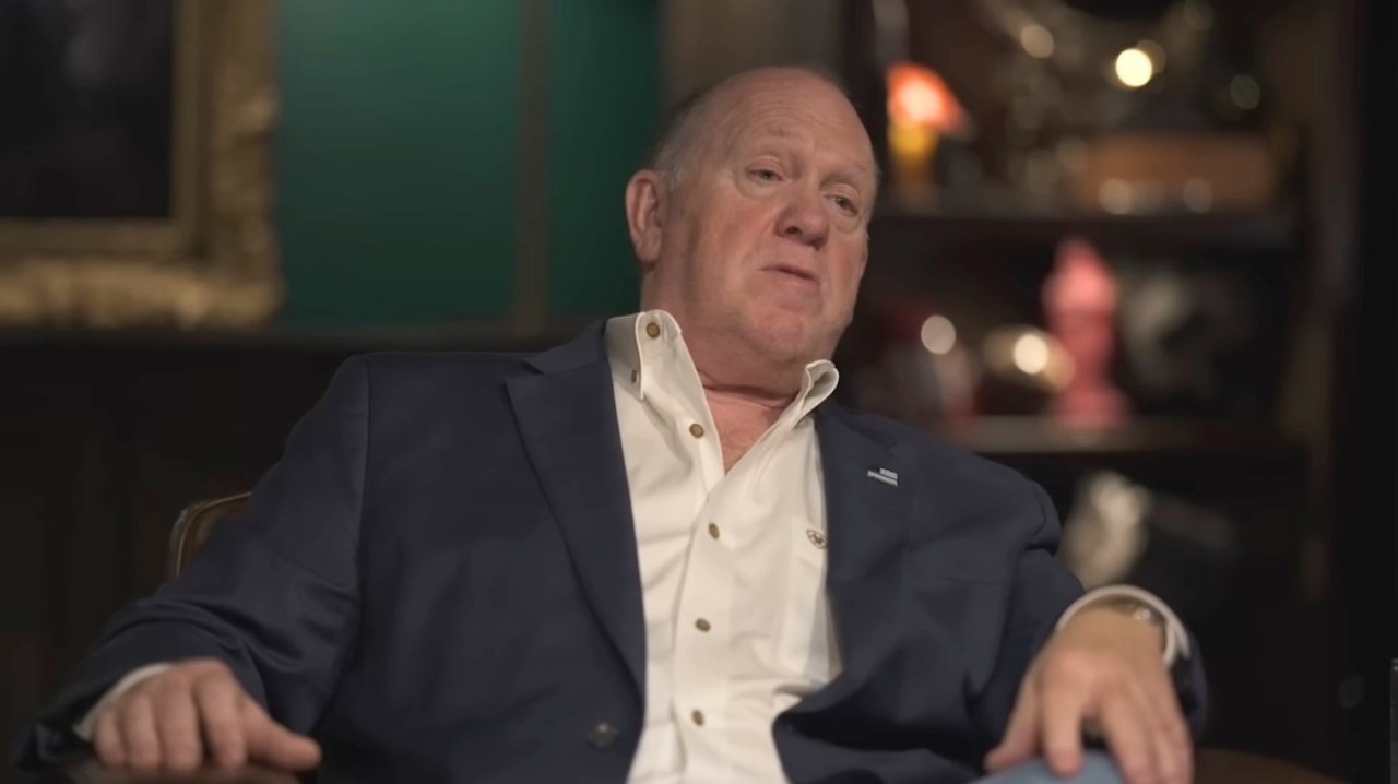 Tom Homan sits in a blue suit and white shirt sits in a dimly lit room