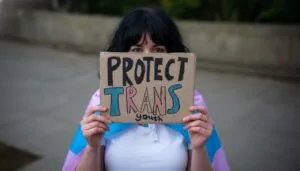 A person holds a cardboard sign reading "Protect Trans Youth" in bold, colorful letters