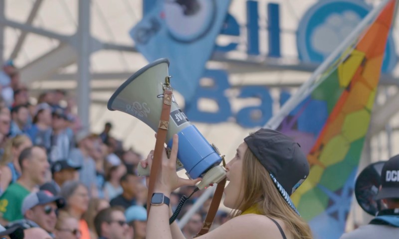 A woman with long hair and a backward cap passionately shouts into a megaphone at a sports event, surrounded by a diverse crowd and colorful flags
