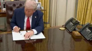 Donald Trump in a suit signs a document at a desk in an official setting