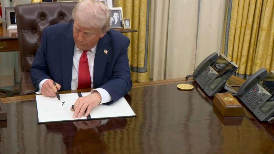 Donald Trump in a suit signs a document at a desk in an official setting