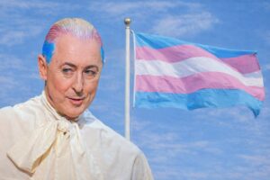 Alan Cumming with blue hair and a white shirt stands in front of a flag