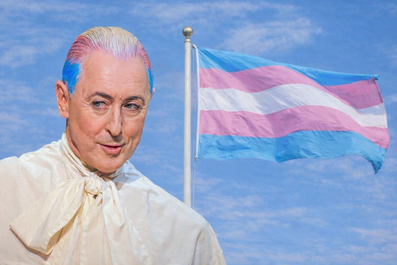 Alan Cumming with blue hair and a white shirt stands in front of a flag
