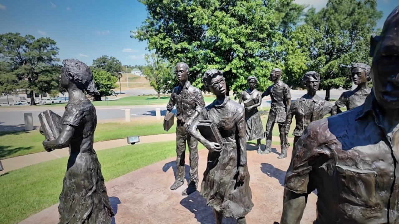Bronze statues of determined men and women holding books stand on a grassy area under a clear blue sky