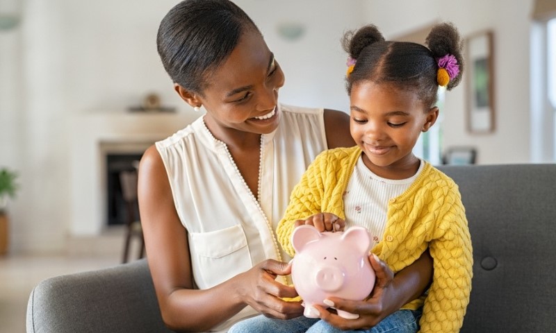 A smiling woman and young girl sit on a couch, holding a pink piggy bank