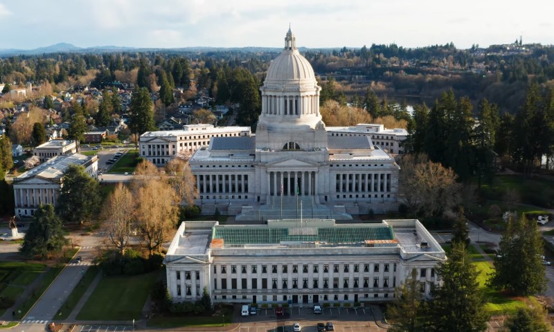 Aerial view of a grand, domed capitol building surrounded by lush trees and adjacent structures, under a partly cloudy sky.