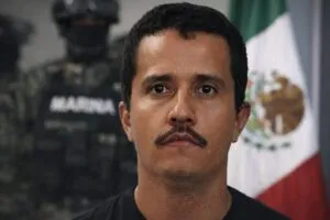 Close-up portrait of El Mencho, with a blurred Mexican flag and security personnel in the background