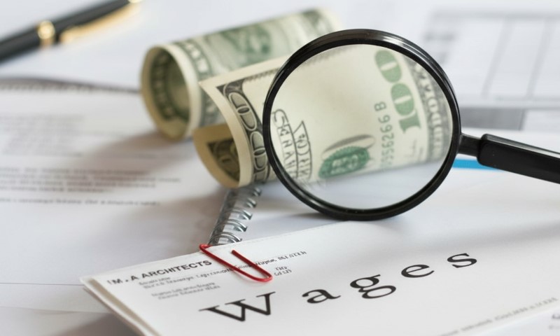A magnifying glass highlights a document titled "Wages," surrounded by rolled-up US dollar bills