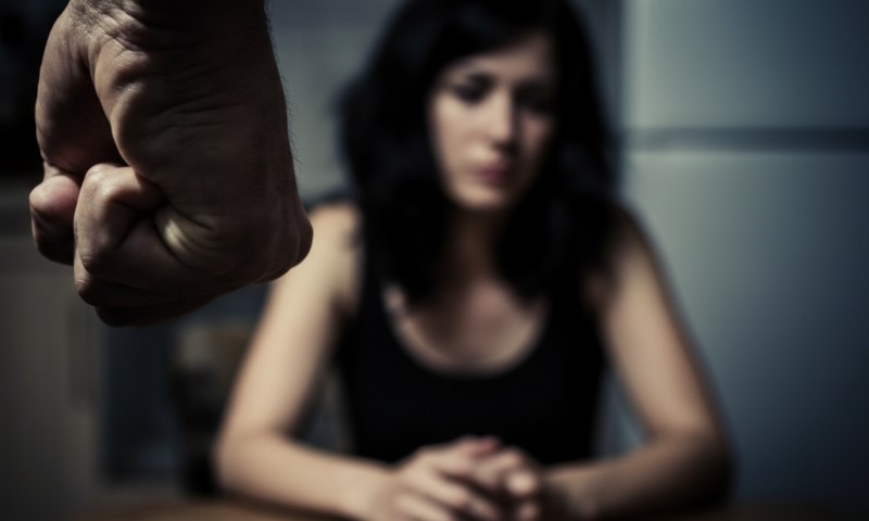 A close-up of a clenched fist in the foreground, with a blurred, seated woman in the background