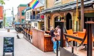 Lively street scene with a couple talking at a café's outdoor bar. A rainbow flag hangs above
