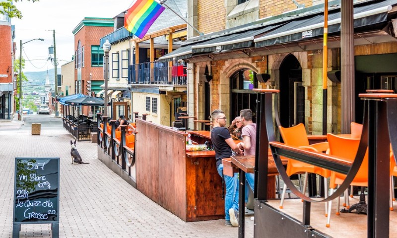 Lively street scene with a couple talking at a café's outdoor bar. A rainbow flag hangs above