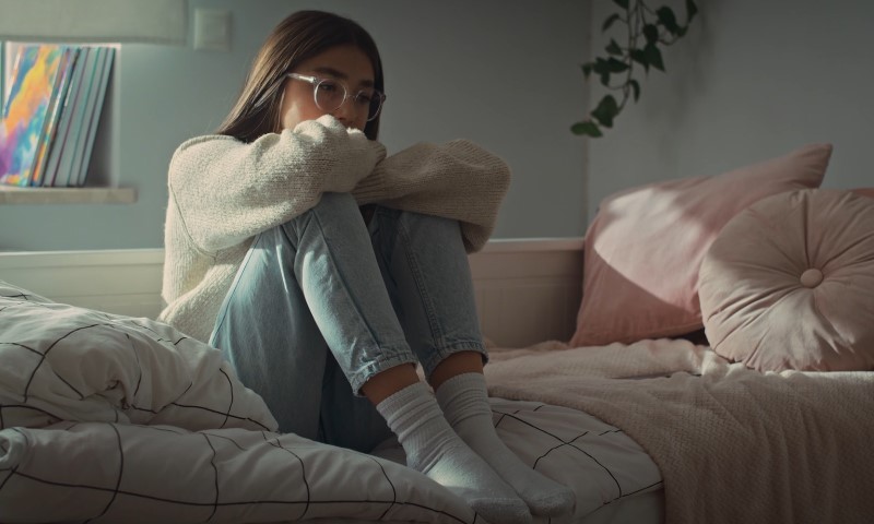 A young woman in glasses sits on a bed, hugging her knees