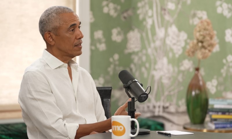 Barack Obama in a white shirt speaks into a microphone, seated at a table with a mug