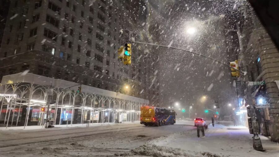 Snowy city street at night with heavy snowfall. A bus and a car drive through snow-covered roads under glowing streetlights. Urban winter scene.