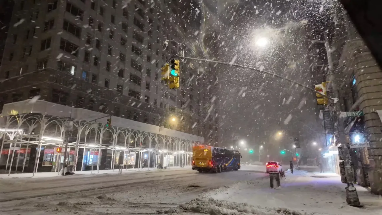 Snowy city street at night with heavy snowfall. A bus and a car drive through snow-covered roads under glowing streetlights. Urban winter scene.