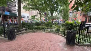 A small brick plaza with two trash bins is surrounded by a black metal fence. Many pride flags line the fence, and trees provide a lush canopy above