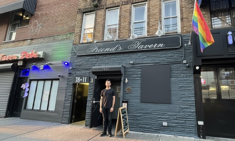 A person stands in front of Friend's Tavern, a building with gray bricks. A rainbow flag hangs above, conveying a welcoming, inclusive atmosphere