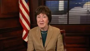 Susan Collins in a tan blazer sits in an office with an American flag behind her