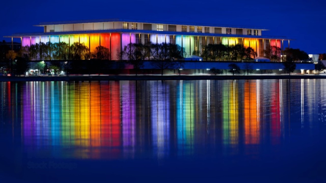 A modern building glows vibrantly with a rainbow of colors at night, reflecting beautifully on the calm water below