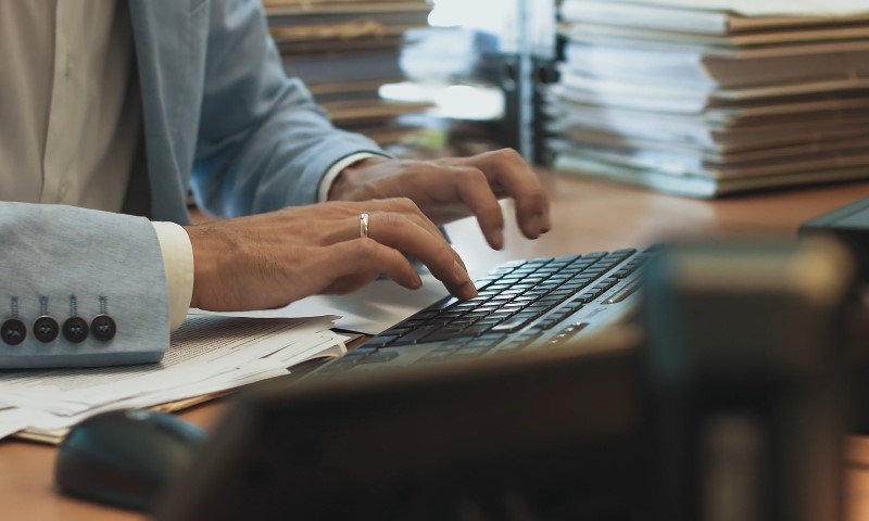 A person in a light blue blazer types on a black keyboard at a cluttered desk with stacks of papers