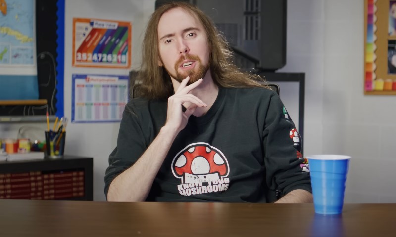 Asmongold with long hair and a beard sits at a table, wearing a dark sweater with a mushroom design