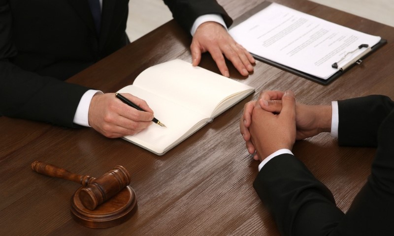 Two people in suits sit at a wooden table, one writing in a notebook and the other with hands clasped