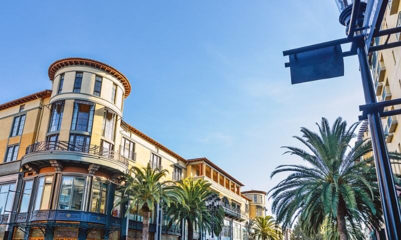 Street view of a vibrant cityscape with Mediterranean-style buildings, tall palm trees, and a bright blue sky