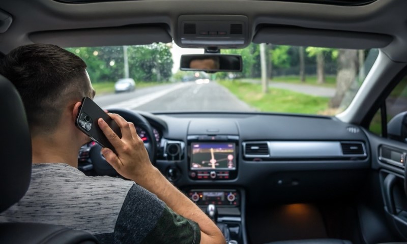 Driver in a car using a smartphone with one hand, road visible through windshield