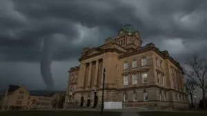 A tornado looms ominously in the dark, cloudy sky near a stately, historic building with a green dome