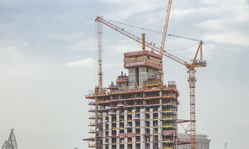 A high-rise building under construction with scaffolding and two cranes against a cloudy sky
