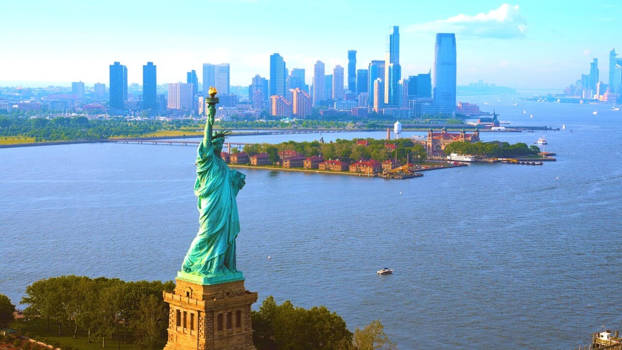 The Statue of Liberty overlooks a calm harbor with boats, lush greenery, and distant skyscrapers under a clear blue sky