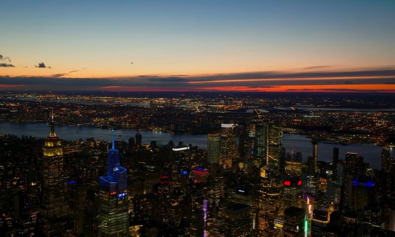 Aerial view of a New York City skyline at dusk, with vibrant building lights below and a colorful sunset above