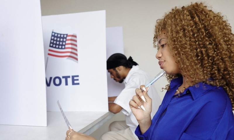 Two people seated at voting booths with privacy panels. A woman in blue is thoughtful, holding a form and pen