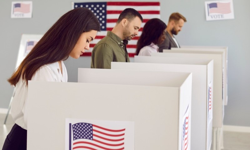 Four people stand at voting booths, casting ballots in a room with American flags