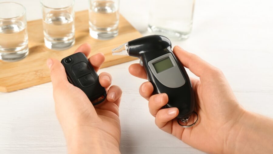 Driver holds a breathalyzer and car key near alcohol glasses, showing challenges in drunk-driving detection technology
