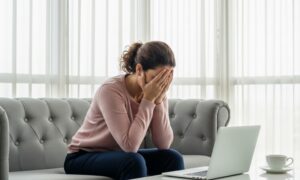 A woman in a pink sweater sits on a gray couch, covering her face with her hands in distress