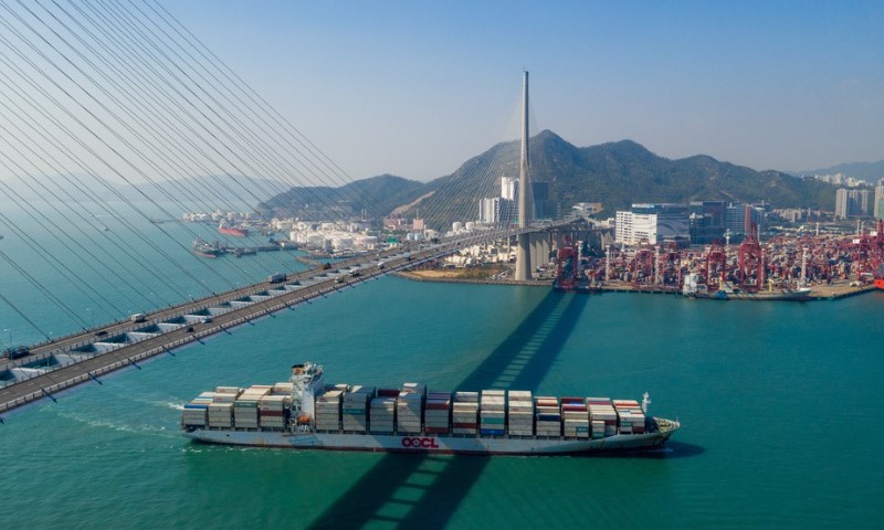 A large container ship sails beneath a suspension bridge in a harbor, with mountains and a busy port in the background