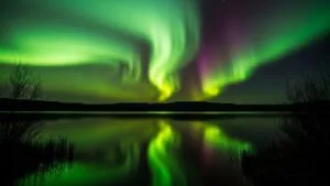 Vivid green and purple northern lights illuminate the night sky, reflecting on a tranquil lake, framed by silhouettes of distant hills and sparse trees