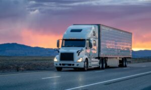 A white semi-truck drives down a highway at sunset, with a dramatic sky in shades of orange and purple